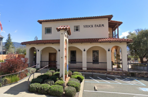 A two-story beige building with a red tile roof and an arched entryway marked "Stock Farm" welcomes both people and dogs. A tall sign stands near the sidewalk. Landscaping includes neatly trimmed bushes along the front. The parking lot has a visible handicap spot close to the entrance, making access easy for all visitors with their pets.